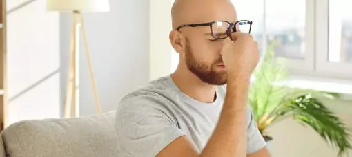 L’homme qui est assis sur le canapé à la maison enlève ses lunettes et se frotte les yeux secs et fatigués. Jeune homme ressentant une douleur dans la région du front, souffrant de tension oculaire, de maux de tête, de migraine oculaire ou de vision floue